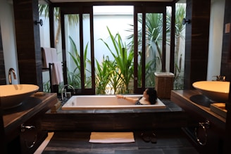 A modern bathroom features a large rectangular bathtub with a person relaxing inside. The room is decorated with sleek, dark wood elements and twin sinks on either side. Large windows offer a view of lush green plants outside, providing a sense of tranquility and connection with nature.