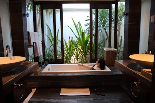 A modern bathroom features a large rectangular bathtub with a person relaxing inside. The room is decorated with sleek, dark wood elements and twin sinks on either side. Large windows offer a view of lush green plants outside, providing a sense of tranquility and connection with nature.