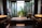 A modern bathroom features a large rectangular bathtub with a person relaxing inside. The room is decorated with sleek, dark wood elements and twin sinks on either side. Large windows offer a view of lush green plants outside, providing a sense of tranquility and connection with nature.