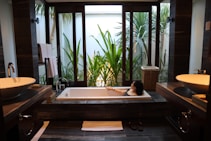 A modern bathroom features a large rectangular bathtub with a person relaxing inside. The room is decorated with sleek, dark wood elements and twin sinks on either side. Large windows offer a view of lush green plants outside, providing a sense of tranquility and connection with nature.
