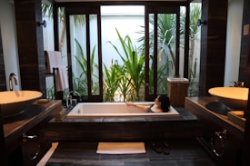 A modern bathroom features a large rectangular bathtub with a person relaxing inside. The room is decorated with sleek, dark wood elements and twin sinks on either side. Large windows offer a view of lush green plants outside, providing a sense of tranquility and connection with nature.