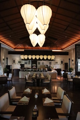 A dimly lit restaurant interior featuring large, lantern-style ceiling lights and neatly arranged dining tables with white napkins. The atmosphere is elegant and serene, with a buffet setup visible at the back. Dark wooden furniture and decor add warmth to the setting, and framed pictures adorn the walls.