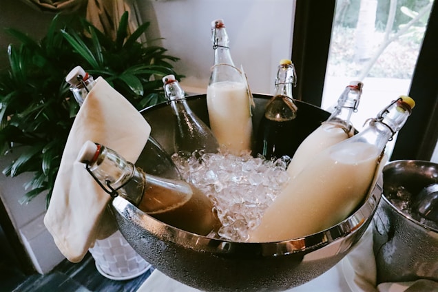 Glass bottles filled with different liquids are placed in a stainless steel bowl filled with ice cubes. The bottles have flip-top caps and are nested among decorative cloths, with a potted plant visible in the background.
