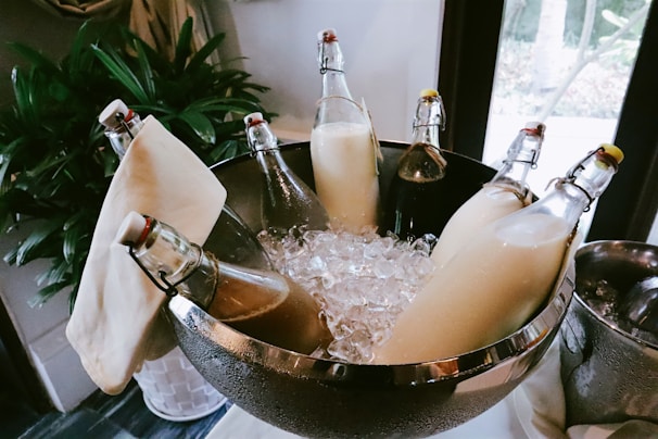 Glass bottles filled with different liquids are placed in a stainless steel bowl filled with ice cubes. The bottles have flip-top caps and are nested among decorative cloths, with a potted plant visible in the background.