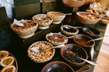 A variety of nuts, seeds, and dried fruits are displayed in woven baskets and bowls on a dark surface. Spoons are placed in each container for serving. The arrangement is visually appealing with an assortment of textures and shapes.