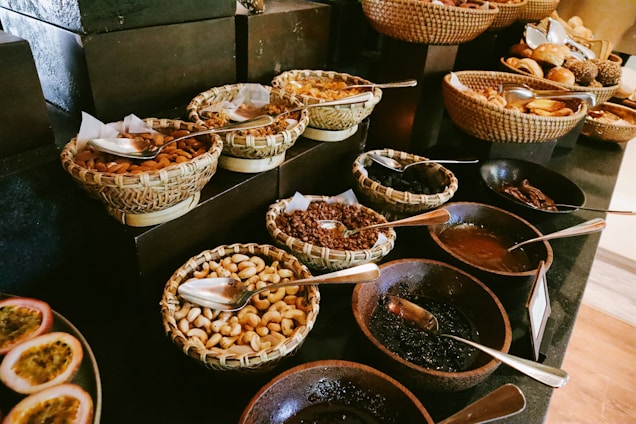 A variety of nuts, seeds, and dried fruits are displayed in woven baskets and bowls on a dark surface. Spoons are placed in each container for serving. The arrangement is visually appealing with an assortment of textures and shapes.