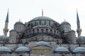 A grand, historic mosque featuring a large central dome surrounded by several smaller domes. Four slender minarets with golden ornaments are visible at the corners. The stone construction is detailed with intricate window patterns, and Arabic script is displayed prominently above the entrance.