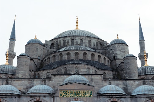 A grand, historic mosque featuring a large central dome surrounded by several smaller domes. Four slender minarets with golden ornaments are visible at the corners. The stone construction is detailed with intricate window patterns, and Arabic script is displayed prominently above the entrance.