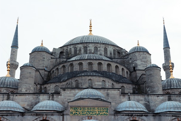 A grand, historic mosque featuring a large central dome surrounded by several smaller domes. Four slender minarets with golden ornaments are visible at the corners. The stone construction is detailed with intricate window patterns, and Arabic script is displayed prominently above the entrance.