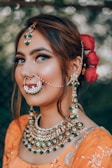A person is wearing traditional jewelry with an intricate nose ring, earrings, and a necklace adorned with green and white jewels. The hair is styled elegantly with red flowers as an accessory. The outfit is a vibrant orange with detailed embroidery.