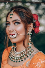 A person is wearing traditional jewelry with an intricate nose ring, earrings, and a necklace adorned with green and white jewels. The hair is styled elegantly with red flowers as an accessory. The outfit is a vibrant orange with detailed embroidery.