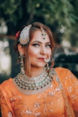 A woman dressed in traditional attire with elaborate jewelry. She is wearing an orange outfit adorned with intricate embroidery and has matching green and gold jewelry, including a large necklace, earrings, and a nose ring. Her makeup is bold, emphasizing her eyes and lips, and her hair is styled elegantly with decorative accessories.