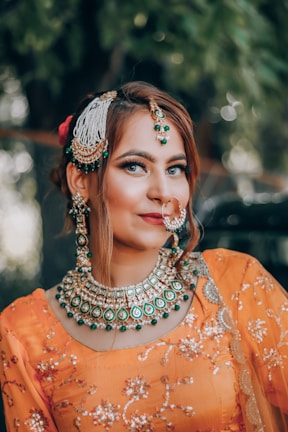 A woman dressed in traditional attire with elaborate jewelry. She is wearing an orange outfit adorned with intricate embroidery and has matching green and gold jewelry, including a large necklace, earrings, and a nose ring. Her makeup is bold, emphasizing her eyes and lips, and her hair is styled elegantly with decorative accessories.