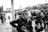 A group of police officers wearing riot gear, including helmets and face shields, positioned on a street. They hold batons and are surrounded by several parked cars and a few bystanders on the sidewalk. The scene appears tense, likely due to the presence of law enforcement in a public area.
