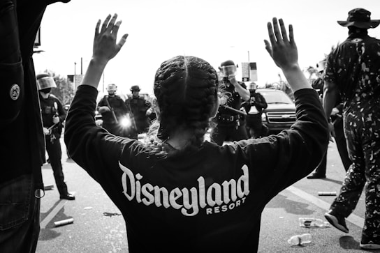 A person with braided hair stands with both arms raised, wearing a sweatshirt with 'Disneyland Resort' printed on the back. A line of law enforcement officers, equipped with helmets and gear, stand in front of the individual. There are scattered objects on the ground, such as bottles and debris. The scene conveys a tense or confrontational atmosphere.