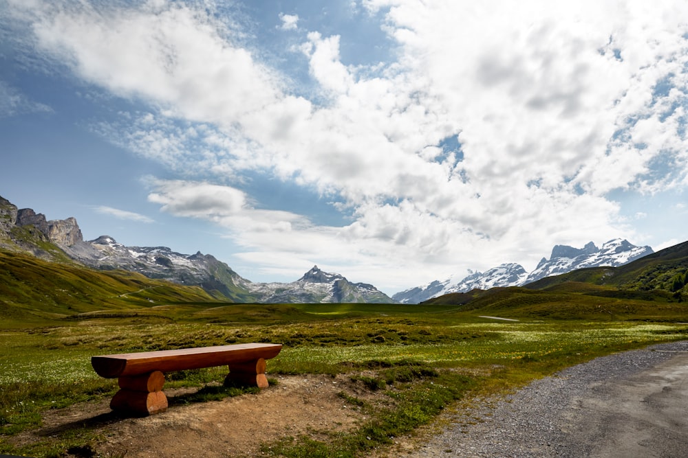 brown wooden bench on green grass field near mountain under white clouds during daytime