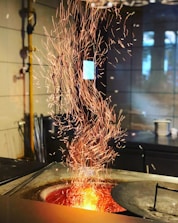 A vibrant display of orange sparks cascading upwards from a glowing furnace. The background features a tiled wall and scattered industrial tools, adding to the workshop ambiance.