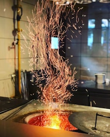 A vibrant display of orange sparks cascading upwards from a glowing furnace. The background features a tiled wall and scattered industrial tools, adding to the workshop ambiance.