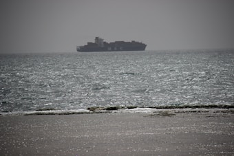 A large cargo ship is sailing on the vast ocean under a hazy sky. The ship is positioned in the middle distance and is partially silhouetted against the bright, shimmering water. The foreground features a stretch of sandy beach with wet areas where the water gently laps against the shore.