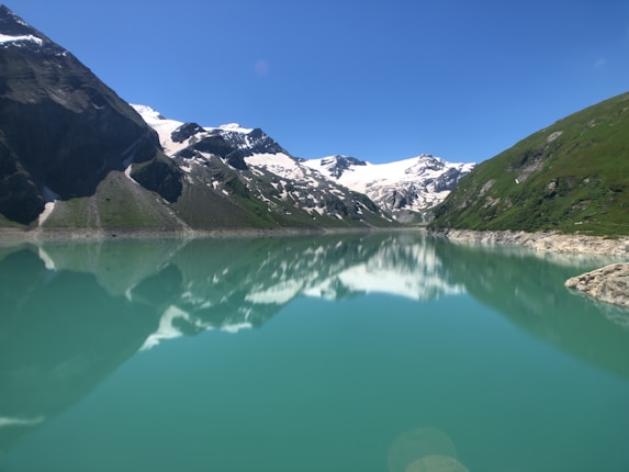 Snow-capped mountains reflected in a tranquil turquoise lake, surrounded by lush green slopes and a clear blue sky.