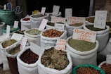 Bags filled with various spices and herbs are displayed in a market setting. Each bag is labeled with handwritten signs in multiple languages, indicating the type of spice or herb. The items are packed in large white fabric sacks, some of which are partially closed or open, revealing the contents like dried herbs, spices, and other dried goods.