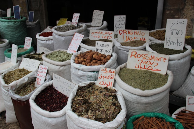 Bags filled with various spices and herbs are displayed in a market setting. Each bag is labeled with handwritten signs in multiple languages, indicating the type of spice or herb. The items are packed in large white fabric sacks, some of which are partially closed or open, revealing the contents like dried herbs, spices, and other dried goods.