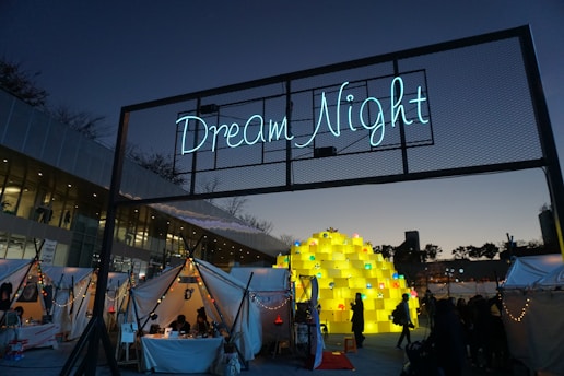 An outdoor festival or event taking place at dusk with a large illuminated sign reading 'Dream Night' displayed above several white tents adorned with string lights. People are gathered around, and a large central sculpture made of stacked yellow blocks, each containing a smaller light or decoration, is prominently featured. The sky is transitioning from day to night, creating a calm atmosphere.