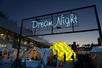 An outdoor festival or event taking place at dusk with a large illuminated sign reading 'Dream Night' displayed above several white tents adorned with string lights. People are gathered around, and a large central sculpture made of stacked yellow blocks, each containing a smaller light or decoration, is prominently featured. The sky is transitioning from day to night, creating a calm atmosphere.
