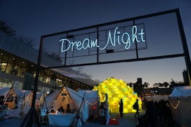 An outdoor festival or event taking place at dusk with a large illuminated sign reading 'Dream Night' displayed above several white tents adorned with string lights. People are gathered around, and a large central sculpture made of stacked yellow blocks, each containing a smaller light or decoration, is prominently featured. The sky is transitioning from day to night, creating a calm atmosphere.