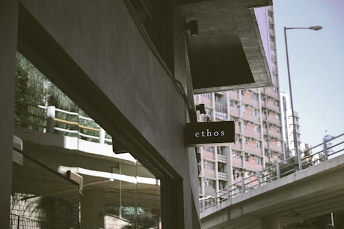 A black sign with the word 'ethos' is mounted on a building. In the background, a tall residential building with several windows is visible, along with an elevated road or bridge with railings. The scene is urban, with muted colors and a somewhat overcast look.
