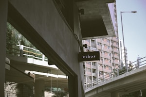 A black sign with the word 'ethos' is mounted on a building. In the background, a tall residential building with several windows is visible, along with an elevated road or bridge with railings. The scene is urban, with muted colors and a somewhat overcast look.