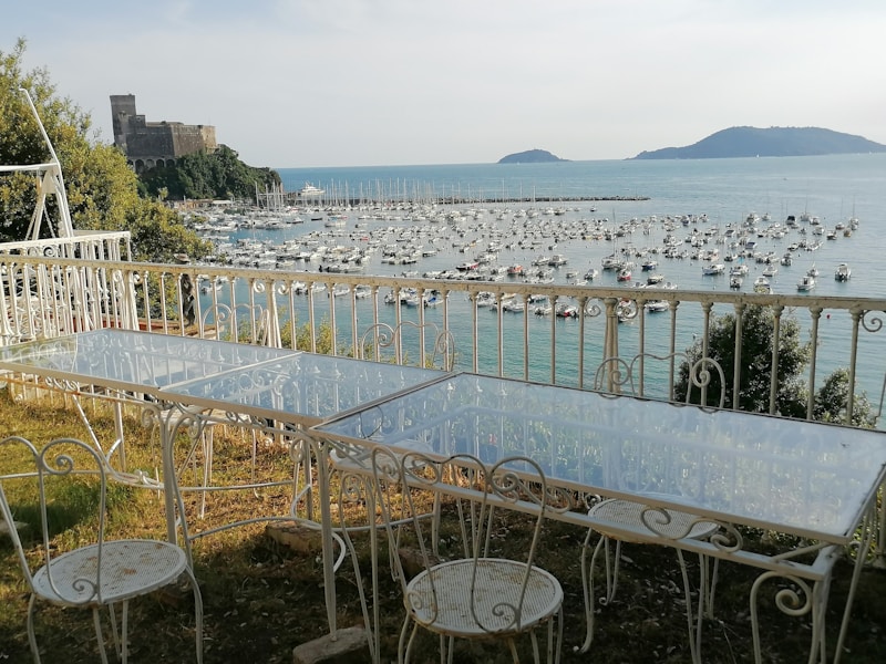 A scenic ocean view from an elevated terrace featuring white wrought iron furniture. Numerous boats are anchored in the harbor, and a castle is visible on a hill to the left. The landscape includes lush greenery and two distant islands on the horizon.