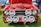 A classic red Ford car with its hood open is on display, showing a detailed view of the engine. The front features multiple large Hella spotlights and a visible license plate with 'PJI 3708'. The car is parked outdoors on grass, with parts of other cars and a person in jeans also visible in the background.