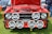 A classic red Ford car with its hood open is on display, showing a detailed view of the engine. The front features multiple large Hella spotlights and a visible license plate with 'PJI 3708'. The car is parked outdoors on grass, with parts of other cars and a person in jeans also visible in the background.