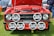 A classic red Ford car with its hood open is on display, showing a detailed view of the engine. The front features multiple large Hella spotlights and a visible license plate with 'PJI 3708'. The car is parked outdoors on grass, with parts of other cars and a person in jeans also visible in the background.