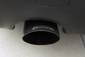 Close-up of a black exhaust pipe with a textured surface and visible branding. The pipe is attached to a vehicle, and the surface appears to have a matte finish. The background is blurred, focusing attention on the exhaust detail.