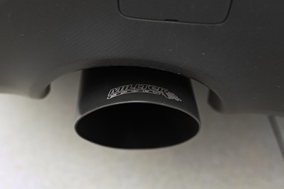 Close-up of a black exhaust pipe with a textured surface and visible branding. The pipe is attached to a vehicle, and the surface appears to have a matte finish. The background is blurred, focusing attention on the exhaust detail.