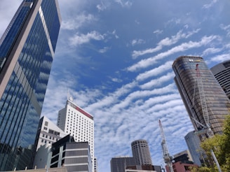Skyscrapers rise against a backdrop of a blue sky filled with patterned clouds. The buildings are modern, with glass facades reflecting the sunlight. Construction cranes are visible, suggesting ongoing urban development.