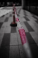 A close-up view of a red and white striped barrier tape stretched across a tiled floor, with the focus primarily on the tape. In the background, a blurred traffic cone is visible.