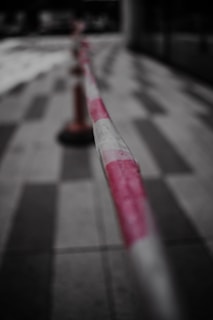 A close-up view of a red and white striped barrier tape stretched across a tiled floor, with the focus primarily on the tape. In the background, a blurred traffic cone is visible.