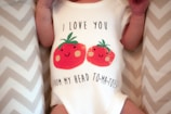A baby is wearing a white onesie featuring a playful design of two smiling red tomatoes with green leaves. Above and below the tomatoes, there is a humorous phrase: 'I love you from my head to-ma-toes.' The background consists of a soft zigzag patterned fabric.