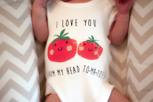 A baby is wearing a white onesie featuring a playful design of two smiling red tomatoes with green leaves. Above and below the tomatoes, there is a humorous phrase: 'I love you from my head to-ma-toes.' The background consists of a soft zigzag patterned fabric.
