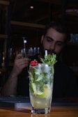 A tall glass filled with a refreshing cocktail garnished with lime slices, mint leaves, and a red flower sits on a wooden table. In the background, a person is preparing a drink, holding a metal bar tool in a dimly lit setting.