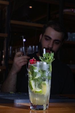 A tall glass filled with a refreshing cocktail garnished with lime slices, mint leaves, and a red flower sits on a wooden table. In the background, a person is preparing a drink, holding a metal bar tool in a dimly lit setting.