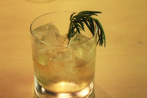 Hand pouring a sparkling cocktail into a tall glass with ice and a sprig of rosemary.