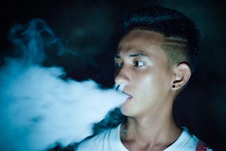 A young person is exhaling a thick cloud of vapor, likely from an electronic cigarette or vape. The background is dark, highlighting the misty smoke as it swirls around.