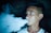 A young person is exhaling a thick cloud of vapor, likely from an electronic cigarette or vape. The background is dark, highlighting the misty smoke as it swirls around.