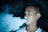 A young person is exhaling a thick cloud of vapor, likely from an electronic cigarette or vape. The background is dark, highlighting the misty smoke as it swirls around.