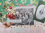 A collage featuring a black-and-white photograph of people in party hats, blowing up balloons and appearing to celebrate. The image is decorated with vintage floral patterns, roses, and cutouts of birds. Text is also included, with words like 'GENUSS', 'GLÜCK', and 'FREUDE' in pastel colors.