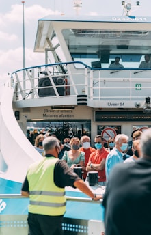 A large ferry with people standing on deck, many wearing face masks. The ferry is labeled 'IJveer 54' and has a prominent railing and control cabin. Bright sunlight casts shadows, and a staff member wearing a high-visibility vest is looking toward the crowd.